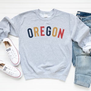 May include: A gray sweatshirt with the word "OREGON" printed in colorful letters. The sweatshirt is paired with blue jeans and white sneakers.