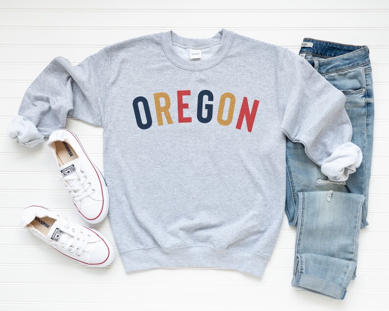May include: A gray sweatshirt with the word "OREGON" printed in colorful letters. The sweatshirt is paired with blue jeans and white sneakers.
