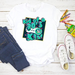 May include: White t-shirt with the text "Kid of a 90's Kid" in a colorful graphic design featuring a retro video game controller. The shirt is paired with blue jeans and white sneakers with red accents. Crayons are also visible.