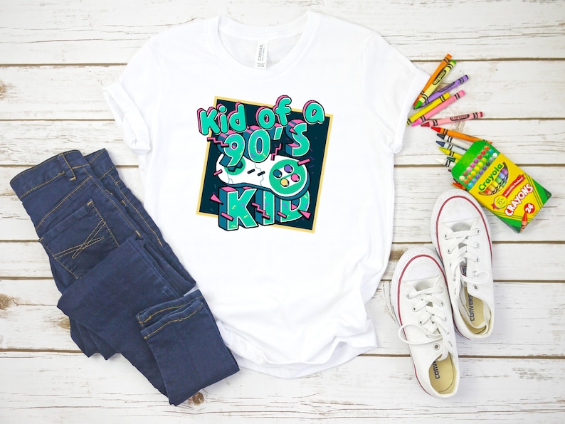 May include: White t-shirt with the text "Kid of a 90's Kid" in a colorful graphic design featuring a retro video game controller. The shirt is paired with blue jeans and white sneakers with red accents. Crayons are also visible.