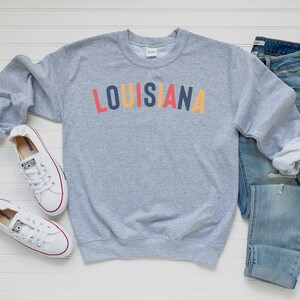 May include: A gray sweatshirt with the word "LOUISIANA" printed in colorful letters. The sweatshirt is paired with blue jeans and white sneakers.