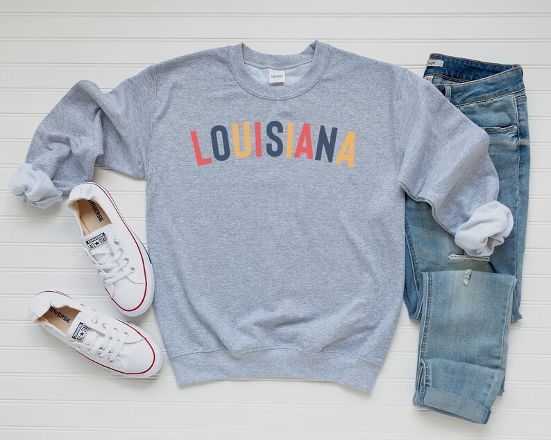 May include: A gray sweatshirt with the word "LOUISIANA" printed in colorful letters. The sweatshirt is paired with blue jeans and white sneakers.