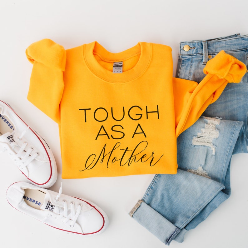 May include: A yellow sweatshirt with the text "TOUGH AS A MOTHER" in black lettering. The sweatshirt is folded and laid on a white surface with a pair of white sneakers and blue jeans.