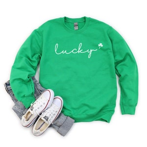 以下が含まれることがあります： 緑色のスウェットシャツに、白い筆記体で「lucky」と書いてあり、その横に小さなクローバーがあります。