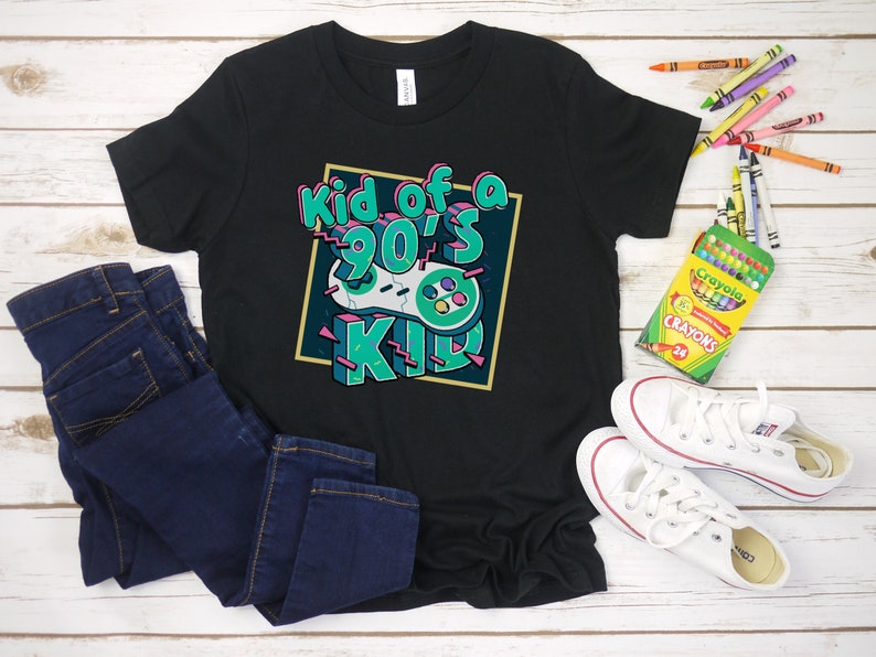 May include: Black t-shirt with the text "Kid of a 90's Kid" in a colorful retro design. The shirt is paired with dark blue jeans and white sneakers. Crayons are scattered around the shirt.