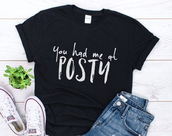 May include: Black t-shirt with the phrase "You had me at POSTY" in white script. The shirt is styled with white sneakers, blue jeans, and a small potted plant. A casual, everyday wear item.
