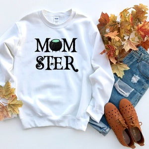 May include: White sweatshirt with black text that reads "MOMSTER" with a black cauldron and spider graphic. The sweatshirt is laying on a white surface with fall leaves and blue jeans.