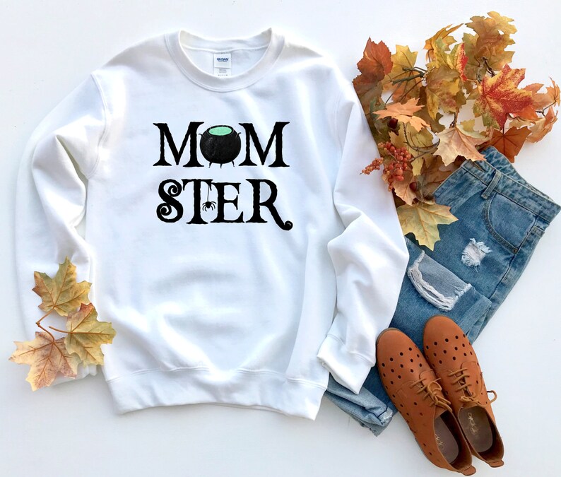 May include: White sweatshirt with black text that reads "MOMSTER" with a black cauldron and spider graphic. The sweatshirt is laying on a white surface with fall leaves and blue jeans.