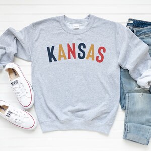 May include: Grey sweatshirt with the word "KANSAS" in colourful block letters. The sweatshirt is paired with blue jeans and white trainers.