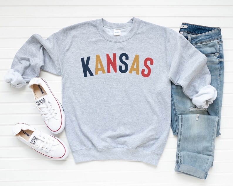 May include: Grey sweatshirt with the word "KANSAS" in colourful block letters. The sweatshirt is paired with blue jeans and white trainers.