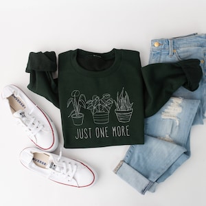 May include: A dark green sweatshirt with a white outline of three potted plants and the text "Just One More". The sweatshirt is laid out on a white surface with a pair of white sneakers and a pair of blue jeans.