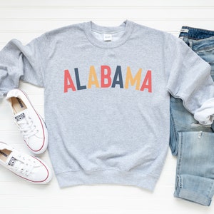 May include: A gray sweatshirt with the word "ALABAMA" printed in red, yellow, and blue letters. The sweatshirt is laid flat on a white surface with a pair of white sneakers and blue jeans.