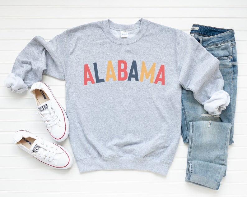May include: A gray sweatshirt with the word "ALABAMA" printed in red, yellow, and blue letters. The sweatshirt is laid flat on a white surface with a pair of white sneakers and blue jeans.