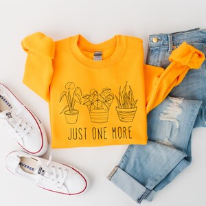 May include: A golden yellow crewneck sweatshirt with a black line drawing of three potted plants and the text "JUST ONE MORE." The sweatshirt is paired with light blue jeans and white canvas sneakers.