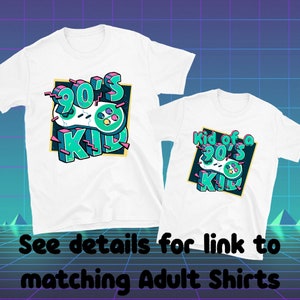 May include: Two white t-shirts with a retro video game controller design. The shirts feature the text "90's Kid" and "Kid of a 90's Kid" in teal, pink, and green. The design is set against a black and gold background.