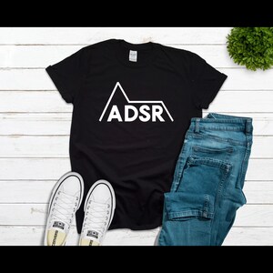 May include: Black t-shirt with a white graphic of a mountain with the word "ADSR" inside. The t-shirt is laying on a white wooden surface with a pair of white sneakers and blue jeans.