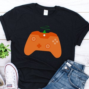 Op de afbeelding: Een zwart T-shirt met een afbeelding van een oranje videogamecontroller in de vorm van een pompoen. De controller heeft een groene steel en een krul bovenop.