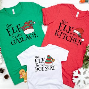 May include: Three Christmas-themed t-shirts in green, red, and white. Each shirt features a playful elf hat graphic and the phrases "the ELF in the GARAGE", "the ELF in the KITCHEN", and "the ELF in the HOT SEAT" in black text.