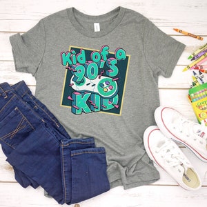 May include: A gray t-shirt with the text "Kid of a 90's Kid" and a video game controller graphic. The shirt is paired with dark blue jeans and white sneakers. Crayons are scattered around the items.