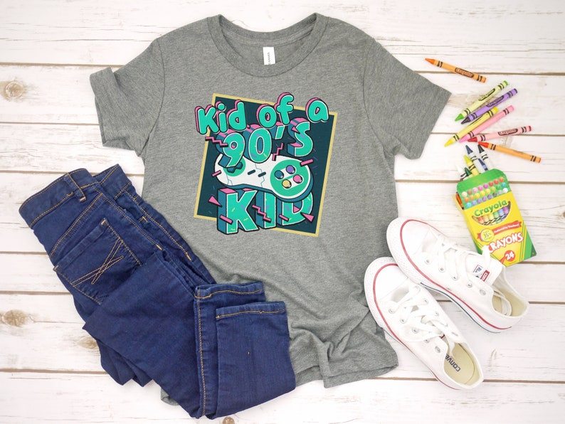 May include: A gray t-shirt with the text "Kid of a 90's Kid" and a video game controller graphic. The shirt is paired with dark blue jeans and white sneakers. Crayons are scattered around the items.