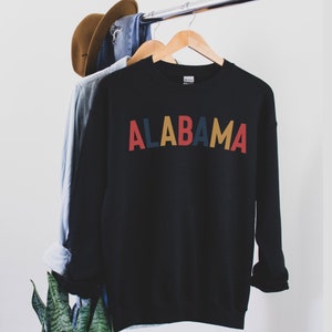 May include: Black sweatshirt with the word "ALABAMA" written in colorful letters. The letters are red, blue, yellow, and red.