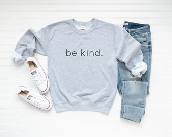 May include: A gray sweatshirt with the text "be kind." printed on the front. The sweatshirt is laid flat on a white surface with a pair of white sneakers and blue jeans.