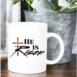 May include: White ceramic mug with the text "He is Risen" in black script and a brown cross. The mug has a curved handle and is set against a blurred background.
