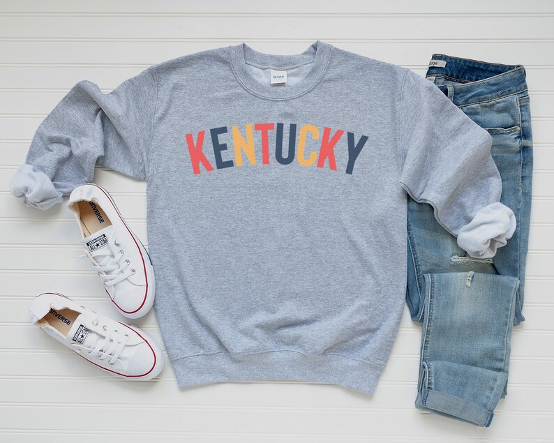 May include: A grey sweatshirt with the word "KENTUCKY" printed in colourful letters. The sweatshirt is paired with blue jeans and white trainers.