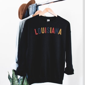 May include: Black sweatshirt with the word "LOUISIANA" printed in colorful letters across the chest.