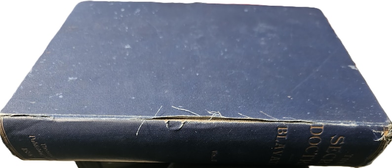 May include: A vintage, hardcover book with a dark blue cover. The spine displays gold lettering and the title "The Secret Doctrine" and the author's name. The book shows signs of wear, with frayed edges and a slightly faded cover.