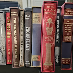 Może przedstawiać: Kolekcja książek w twardej oprawie, w tym "The Maltese Falcon" i "Hans Andersen's Fairy Tales." Grzbiety książek są w kolorach czerwonym, brązowym i niebieskim. Książki pochodzą z wydawnictwa Folio Society.