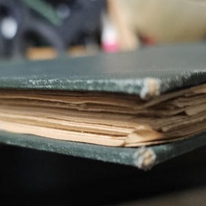Op de afbeelding: Close-up van een oud boek met een donkergroene kaft en vergeelde pagina's. De randen van het boek vertonen slijtage, met licht beschadigde hoeken. De pagina's zijn opgestapeld, wat de dikte van het boek aantoont.