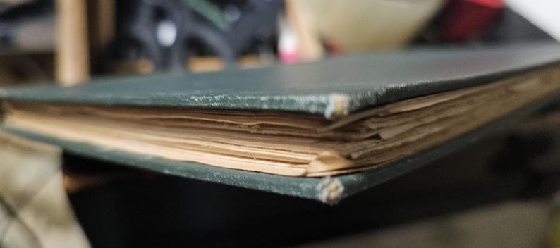 Op de afbeelding: Close-up van een oud boek met een donkergroene kaft en vergeelde pagina's. De randen van het boek vertonen slijtage, met licht beschadigde hoeken. De pagina's zijn opgestapeld, wat de dikte van het boek aantoont.