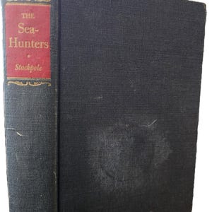 Op de afbeelding: Een hardcover boek getiteld "The Sea-Hunters" van Stackpole. Het boek heeft een donkerblauwe kaft met een rood en goud label op de rug. De kaft vertoont tekenen van slijtage.