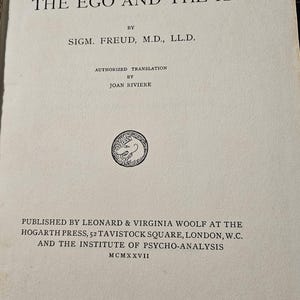 Op de afbeelding: Een vintage boek getiteld "The Ego and the Id" van Sigmund Freud. De omslag bevat zwarte tekst, waaronder de naam van de auteur, de uitgever en het jaar MCMXXVII. Onderdeel van "The International Psycho-Analytical Library".