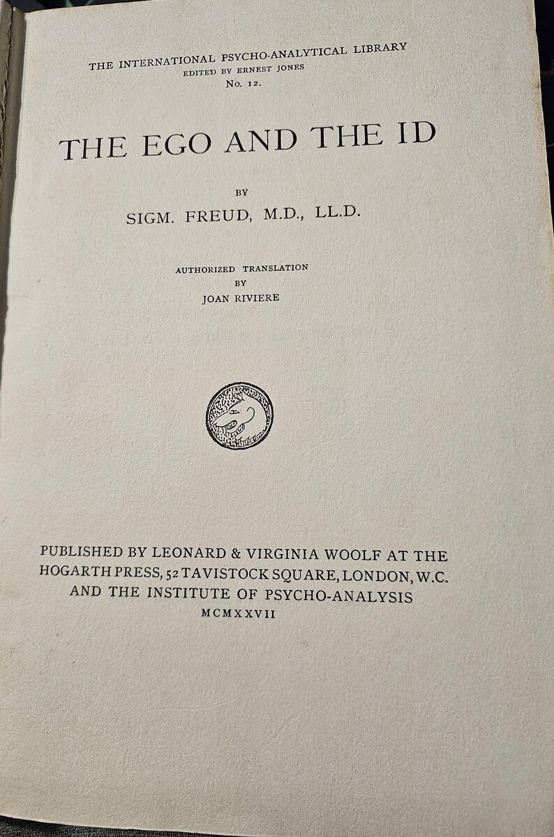 Op de afbeelding: Een vintage boek getiteld "The Ego and the Id" van Sigmund Freud. De omslag bevat zwarte tekst, waaronder de naam van de auteur, de uitgever en het jaar MCMXXVII. Onderdeel van "The International Psycho-Analytical Library".