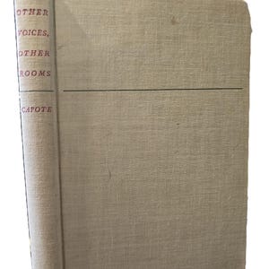 May include: A vintage hardback book with a beige linen cover. The title "Other Voices, Other Rooms" by Truman Capote is printed in red on the spine.