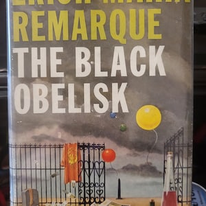 May include: A book cover with the title "The Black Obelisk" by Erich Maria Remarque. The cover features a black metal gate with a red and yellow cloth hanging from it. There are three balloons in the sky, one yellow, one red, and one blue. The background is a cloudy sky.