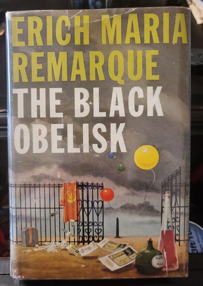 May include: A book cover with the title "The Black Obelisk" by Erich Maria Remarque. The cover features a black metal gate with a red and yellow cloth hanging from it. There are three balloons in the sky, one yellow, one red, and one blue. The background is a cloudy sky.