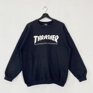 Pode incluir: Camisola preta de gola redonda com o texto branco "Thrasher" acima de "Skateboard Magazine". A camisola está pendurada num cabide preto sobre um fundo branco. A camisola tem mangas compridas e decote canelado.