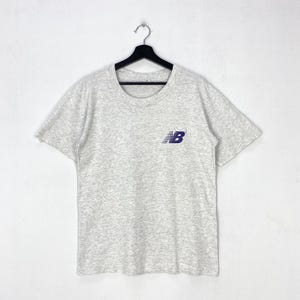 May include: A light gray heathered short-sleeve t-shirt featuring a dark blue New Balance logo on the left chest. The garment is displayed on a black plastic hanger against a plain white background, showcasing a crew neckline and a standard fit.
