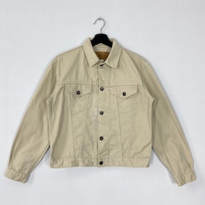 May include: A tan denim jacket with a classic collar, button-down front, and two chest pockets. The jacket has a small red tag on the left chest pocket and is hanging on a black hanger.