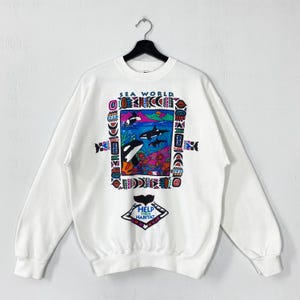 May include: White sweatshirt with a colourful graphic of orcas in the ocean, framed by geometric shapes. The text "SEA WORLD" is at the top, and "HELP THEIR HABITAT" is at the bottom.