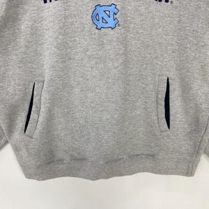May include: A gray hoodie with a front pocket and a light blue logo. The logo features the letters "NC" in a stylized design. The hoodie has a drawstring hood and long sleeves. The fabric appears to be a soft, comfortable material.
