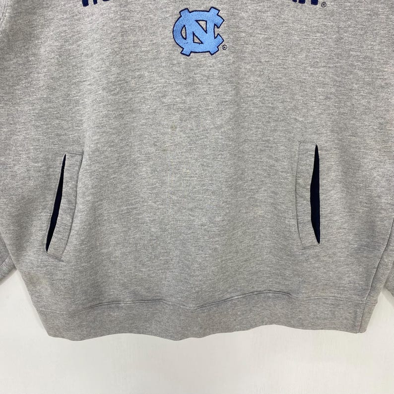 May include: A gray hoodie with a front pocket and a light blue logo. The logo features the letters "NC" in a stylized design. The hoodie has a drawstring hood and long sleeves. The fabric appears to be a soft, comfortable material.