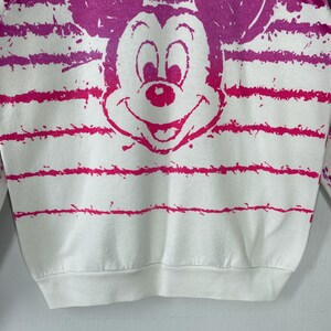 May include: White sweatshirt featuring a large, pink Mickey Mouse graphic. The design includes horizontal pink stripes and splatters. The sweatshirt has a crew neck and long sleeves, perfect for casual wear.