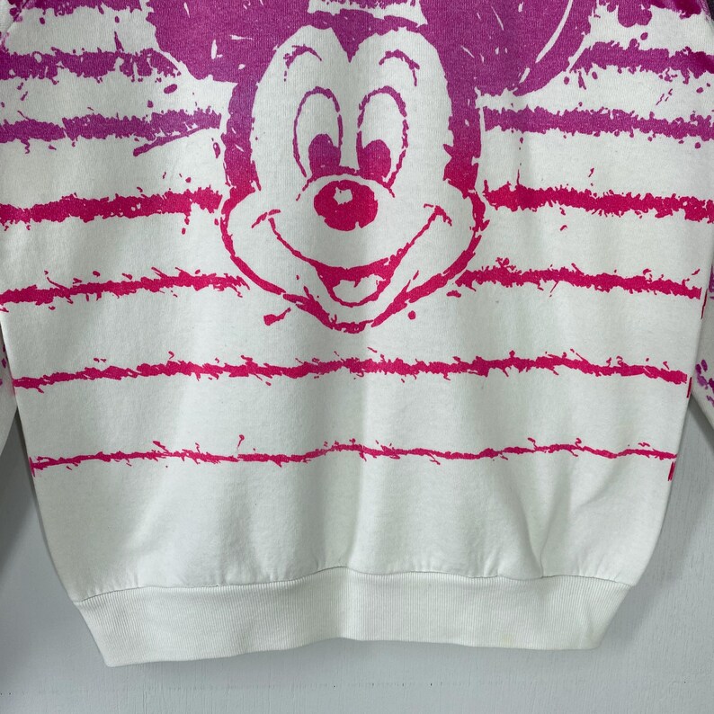 May include: White sweatshirt featuring a large, pink Mickey Mouse graphic. The design includes horizontal pink stripes and splatters. The sweatshirt has a crew neck and long sleeves, perfect for casual wear.