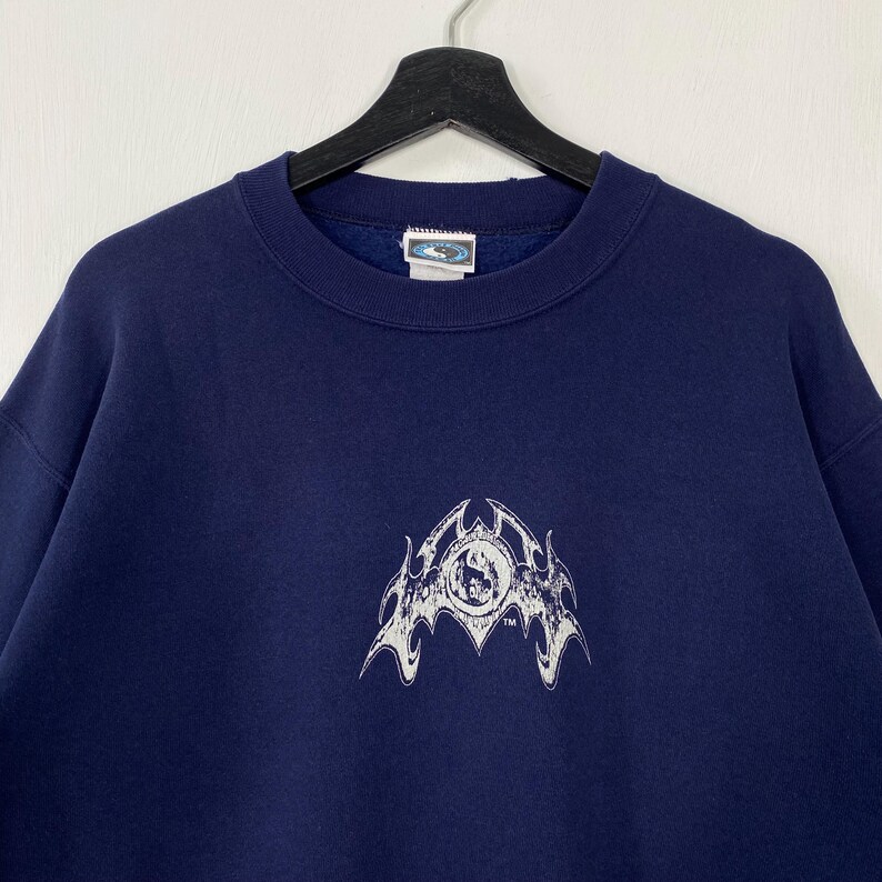 Puede incluir: Sudadera azul marino con un dise&ntilde;o gr&aacute;fico blanco en la parte delantera. El gr&aacute;fico es un emblema estilizado con un centro circular y elementos en forma de llama. La sudadera tiene cuello redondo y mangas largas.