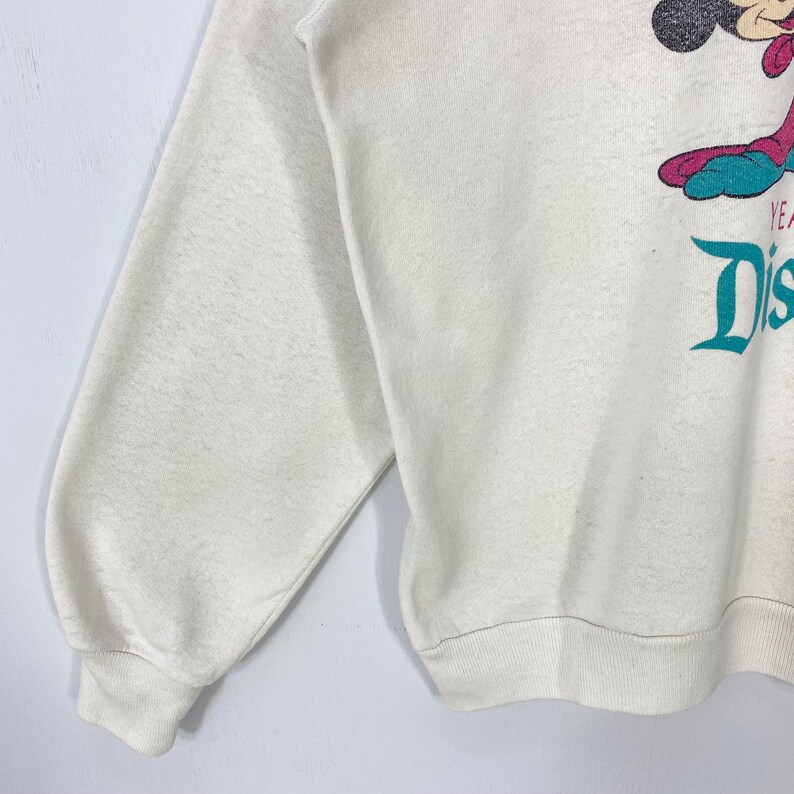 Pu&ograve; includere: Felpa color crema con grafica di Topolino in viola e turchese. Il testo turchese recita "YEARS" e "Dis". La felpa ha maniche lunghe e girocollo.
