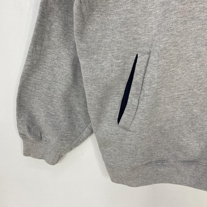 May include: Close-up of a gray sweatshirt with a pocket. The sweatshirt has a dark blue pocket lining. The fabric appears to be a soft, medium-weight material. The sleeve cuff is visible on the left side of the image.